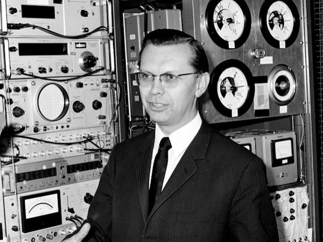 Ein Physiker steht vor technischen Geräten, auf denen Messskalen zu erkennen sind und in die Kabel hinein- und herausführen.