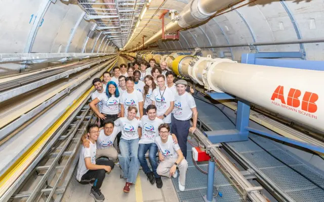 Group of young scientists in T-shirts inside particle accelerator tunnel next to large ABB pipe system.