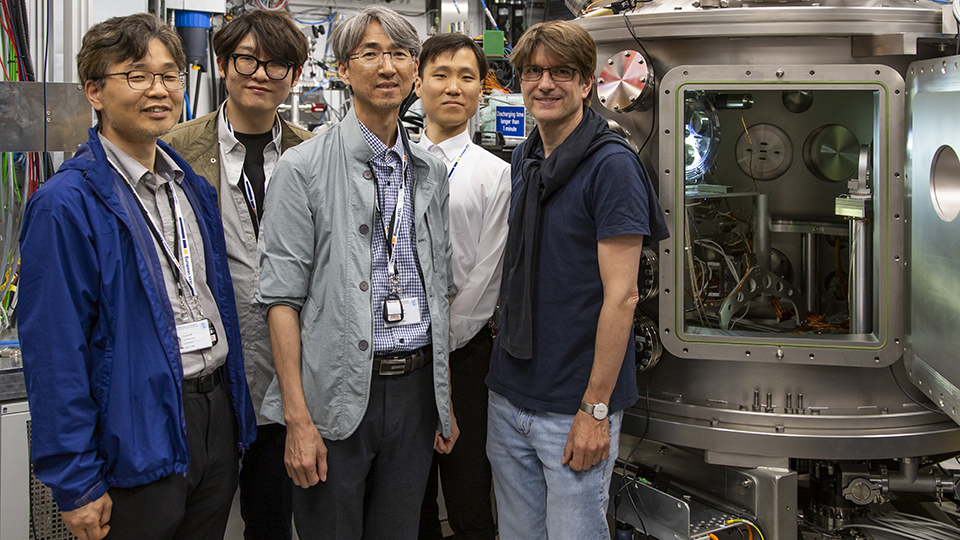 Five men stand smiling in a brightly lit high-tech laboratory, next to them a large metal chamber with its door ajar. Five men stand smiling in a brightly lit high-tech laboratory, next to them a large metal chamber with its door ajar.