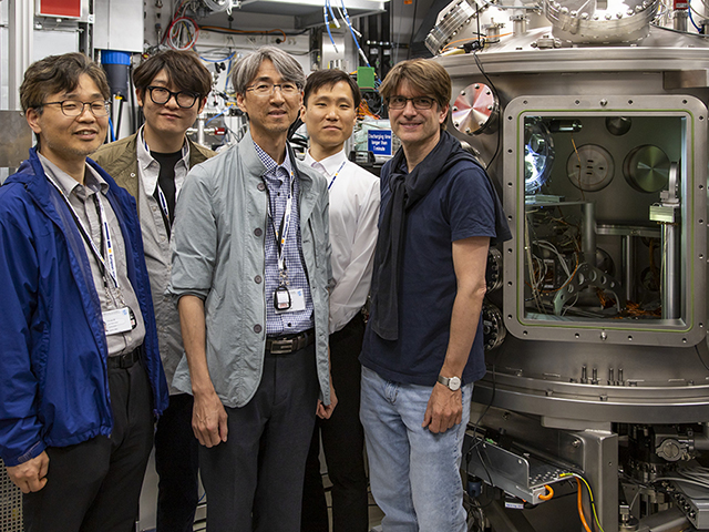 Five men stand smiling in a brightly lit high-tech laboratory, next to them a large metal chamber with its door ajar.
