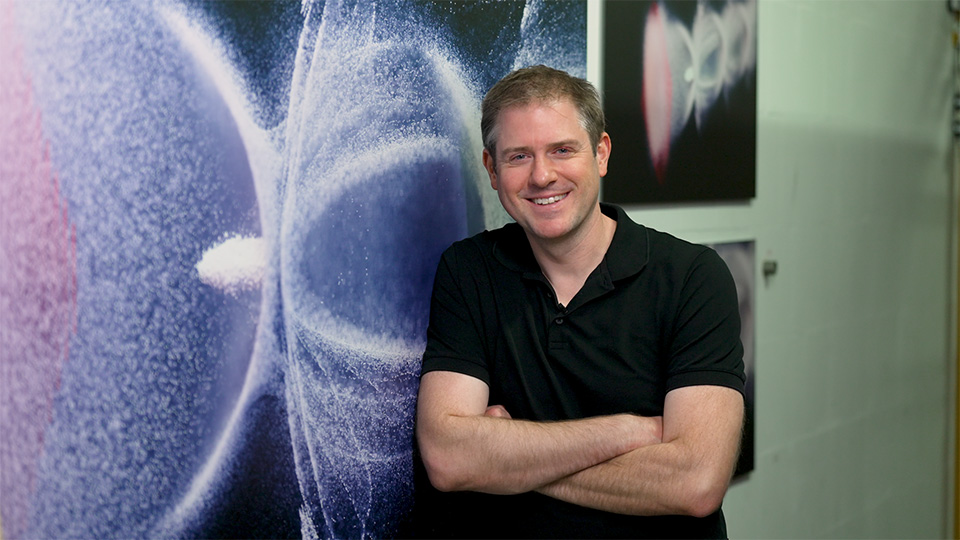 A man stands with crossed arms smiling next to an illustration of accelerated particles.