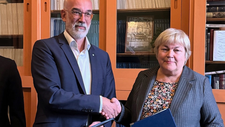 A man and a woman, both in suits and looking serious, shake hands in front of a bookshelf. German, European and Ukranian flags can be seen in the foreground.