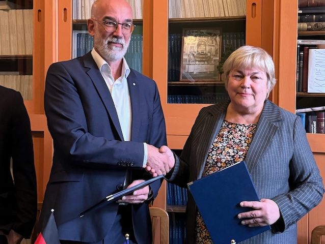 Ein Mann und eine Frau, beide in formeller Kleidung und mit würdevollem Gesichtsausdruck, geben sich vor einem Bücherregal die Hand. Im Vordergrund sind deutsche, europäische und ukrainische Flaggen zu sehen.