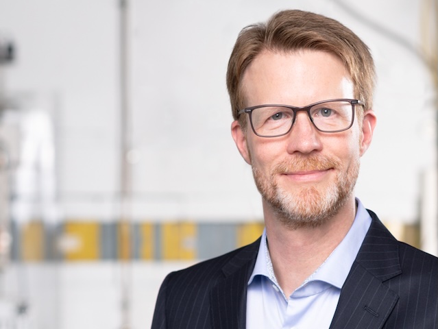 Portrait eines freundlich lächelnden Mannes im blauen Nadelstreifensakko und hellblauem Hemd vor wissenschaftlicher Apparatur