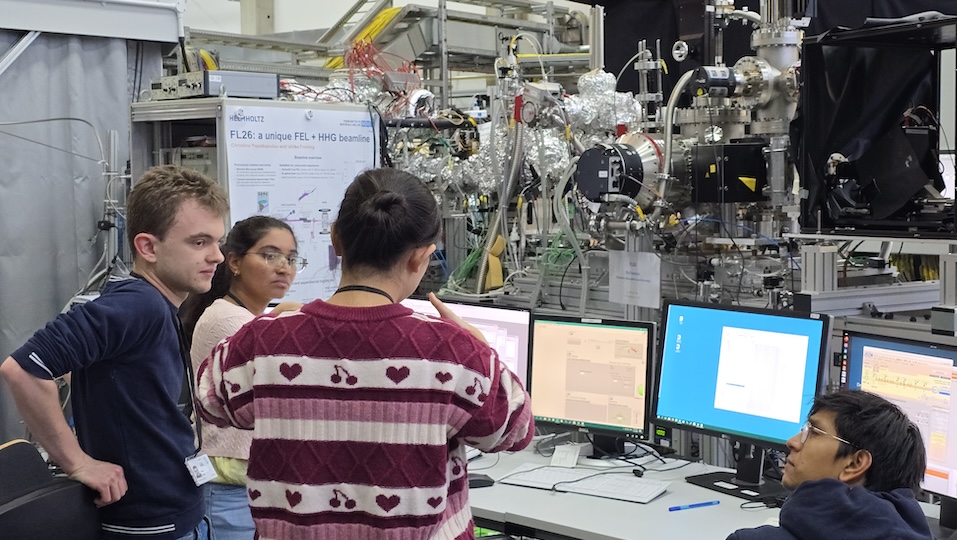 Eine Wissenschaftlerin, die der Kamera den Rücken zudreht, erklärt einer Kollegin und einem Kollegin ein im Hintergrund zu sehenden wissenschaftliches Instrument.