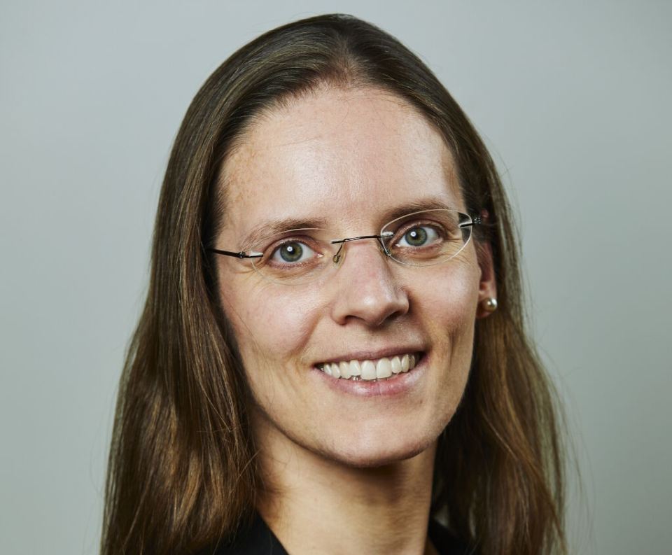 Katharina Behr in a portrait with long brown hair, glasses, and dark clothing against a neutral background.