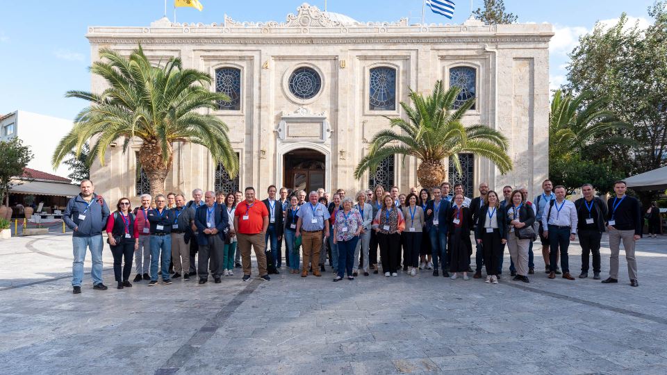 Group of scientists posing in front of historic building with palm trees and flags.