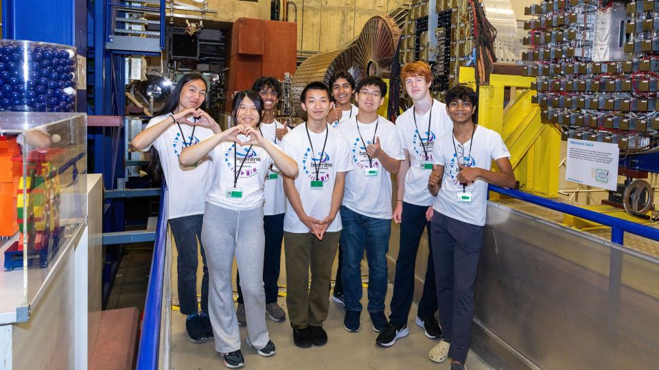 Group of eight teenagers in white T-shirts posing in a technical lab setting with particle accelerator.