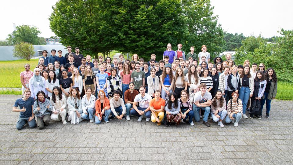 Große Gruppe junger Erwachsener posiert draußen vor Bäumen für ein Gruppenfoto bei sonnigem Wetter.