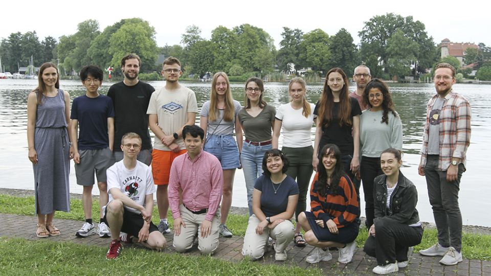Gruppe von 16 jungen Erwachsenen posiert am Ufer eines Sees mit Bäumen und Gebäuden im Hintergrund.