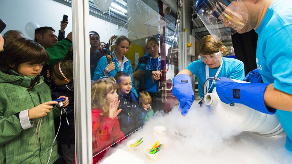 Kinder und Erwachsene beobachten durch eine Glasscheibe, wie zwei Personen in blauen T-Shirts und mit Schutzbrillen eine Demonstration mit flüssigem Stickstoff durchführen.