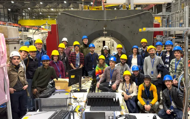 Group of scientists wearing safety helmets posing in front of a large experimental setup in a laboratory hall.