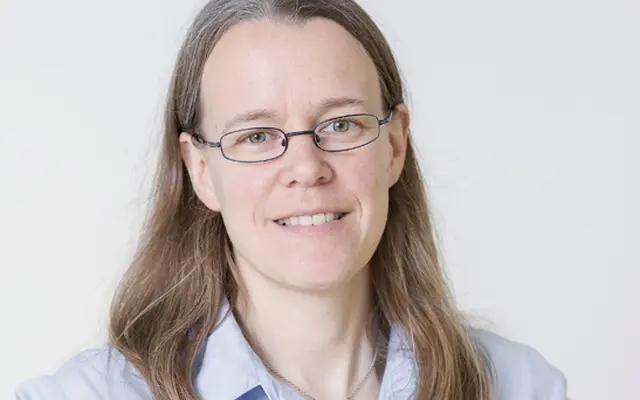 Kerstin Tackmann bei einem Portrait vor hellem Hintergrund, trägt Brille und hellblaues Hemd, schaut freundlich in die Kamera.