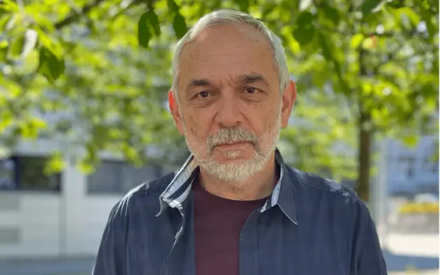 Sergii Fomin bei einem Portrait im Freien vor grünen Bäumen, trägt Hemd und T-Shirt, blickt ruhig in die Kamera.