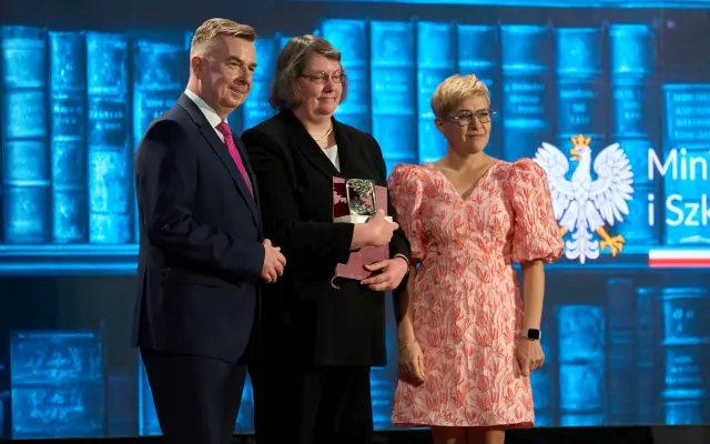 Drei Personen posieren auf einer Bühne, eine hält eine Auszeichnung, im Hintergrund das Logo eines Ministeriums mit Adleremblem.