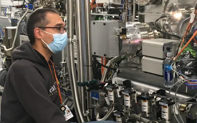 Forschende Person mit Maske arbeitet an einer komplexen wissenschaftlichen Apparatur mit Metallrohren, Kabeln und Messgeräten.