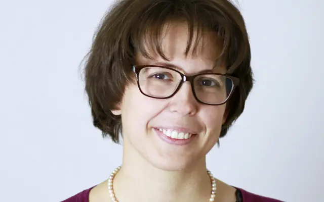 Priscilla Pani bei einem Portrait vor hellem Hintergrund, trägt Brille, Perlenkette und bordeauxrote Strickjacke, lächelt in die Kamera.