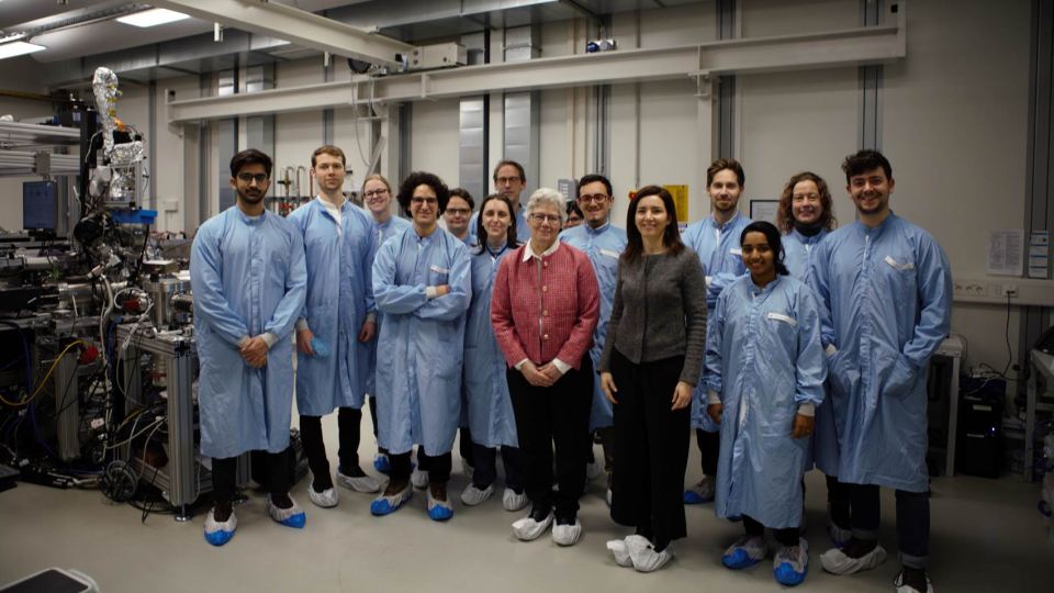 Gruppe von Wissenschaftler:innen in blauer Schutzkleidung in einem Hightech-Labor mit Geräten und Kabeln.