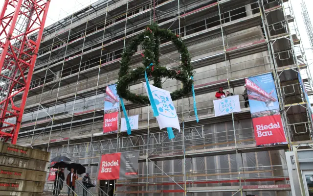 Kranz mit DESY-Flagge hängt am Kran vor einem eingerüsteten Rohbau beim Richtfest, Banner von Riedel Bau sichtbar.