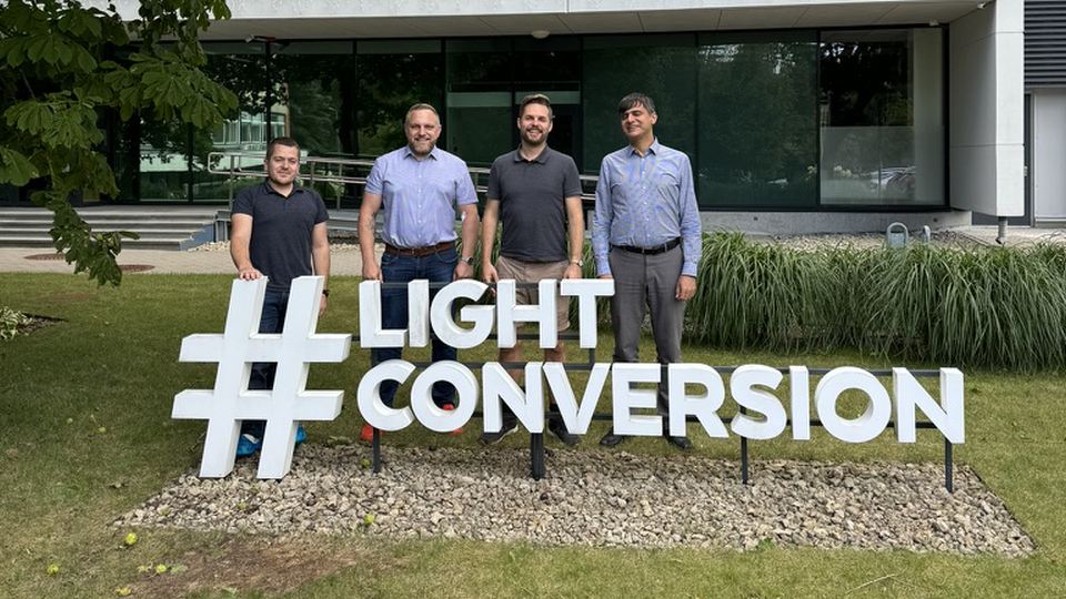 Four people standing by the “#LIGHT CONVERSION” sign outside a modern office building.