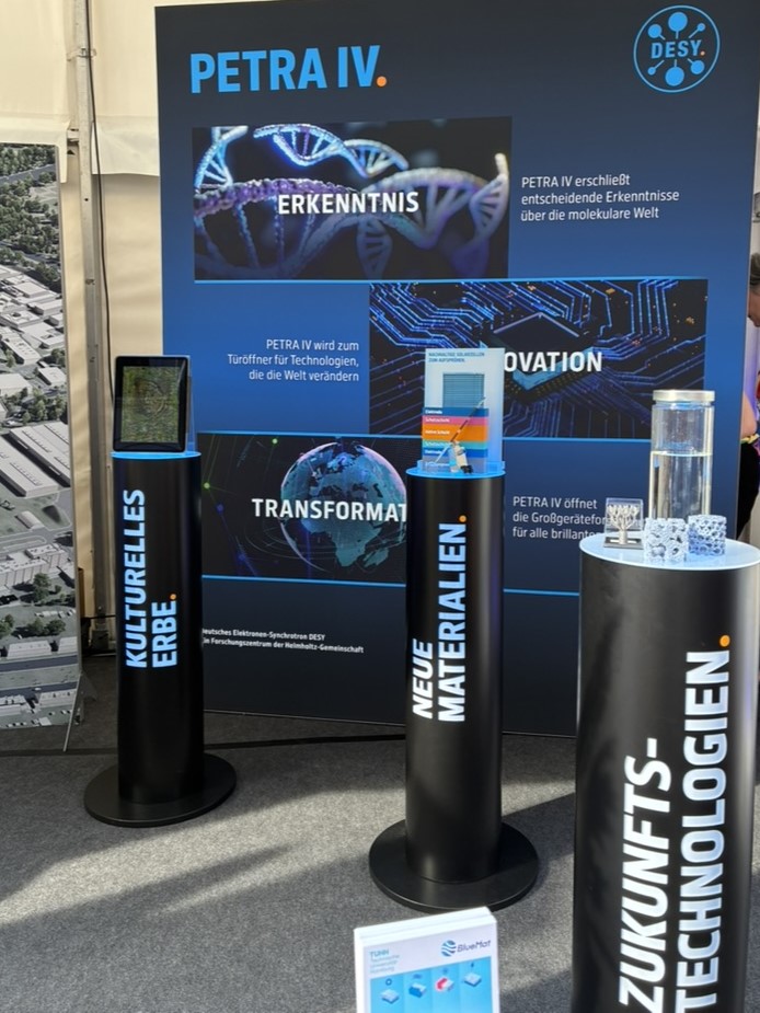 Blick in den Messestand mit Messewand und runden Säulen auf denen Exponate stehen.