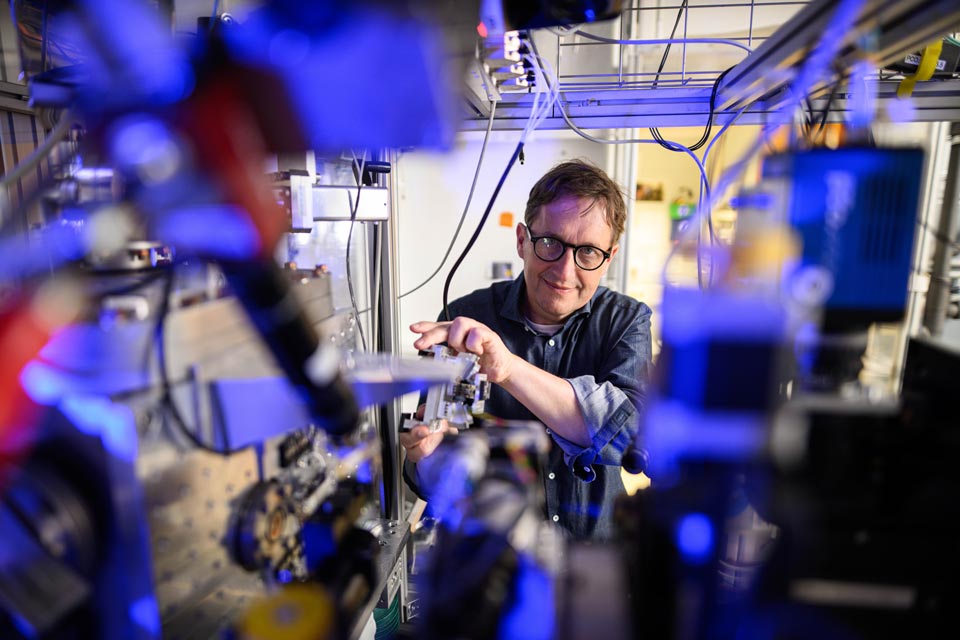 Ein Forscher steht eingerahmt von einem komplexen technischen Aufbau an der Experimentierstation und lächelt zufrieden in die Kamera.